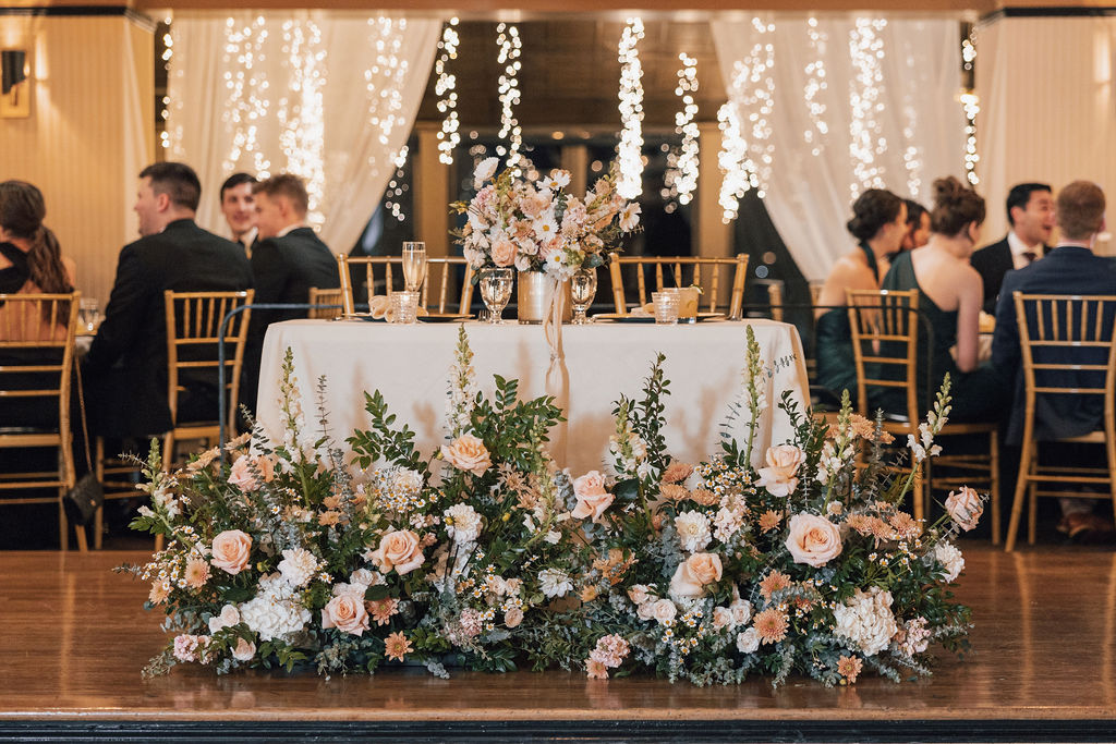 A sweetheart table with pink and white flowers and greenery at the base