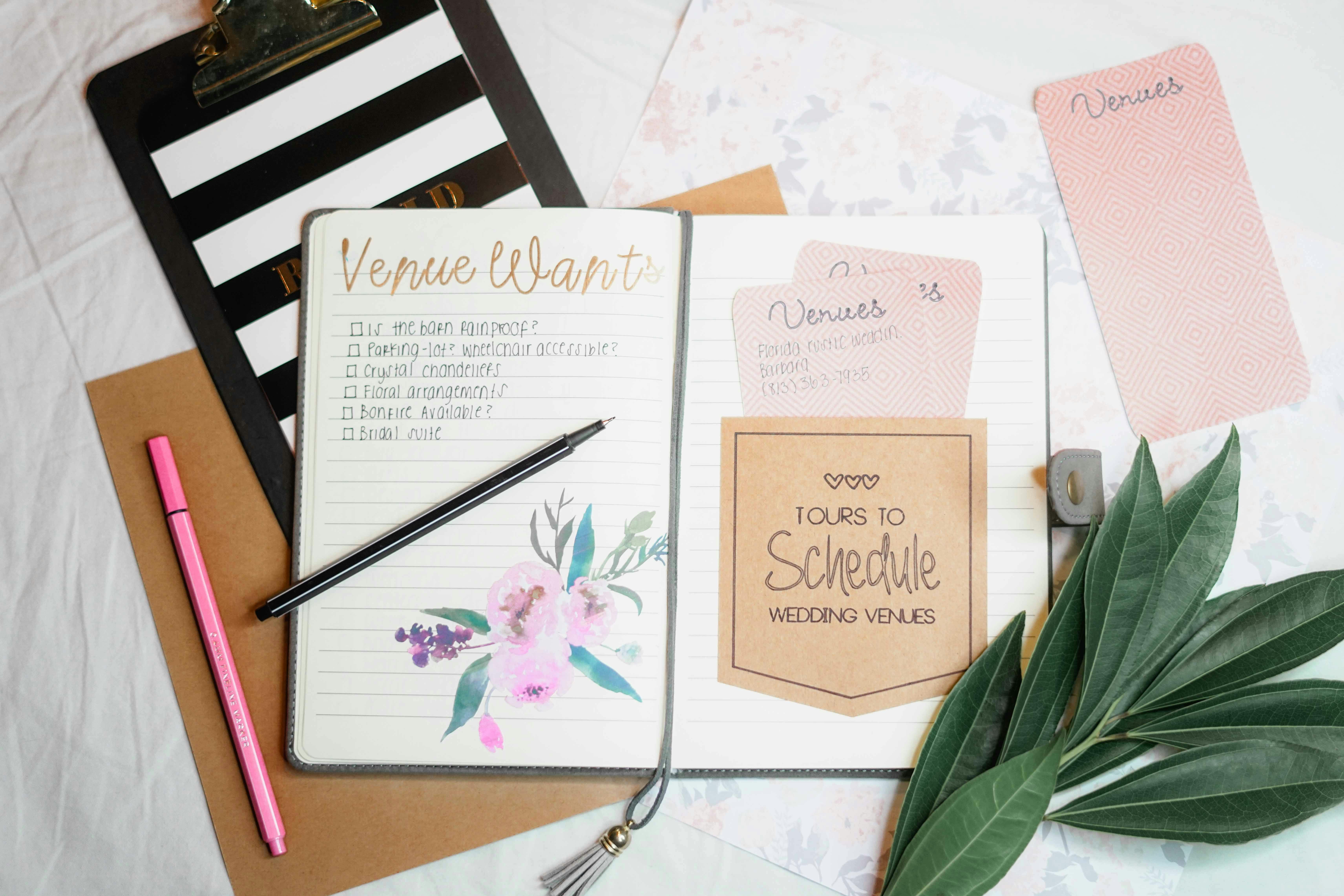 A styled flat lay featuring an open wedding planning notebook with 'Venue Wants' and 'Tours to Schedule' lists, a black and pink pen, a striped clipboard, and decorative venue cards.