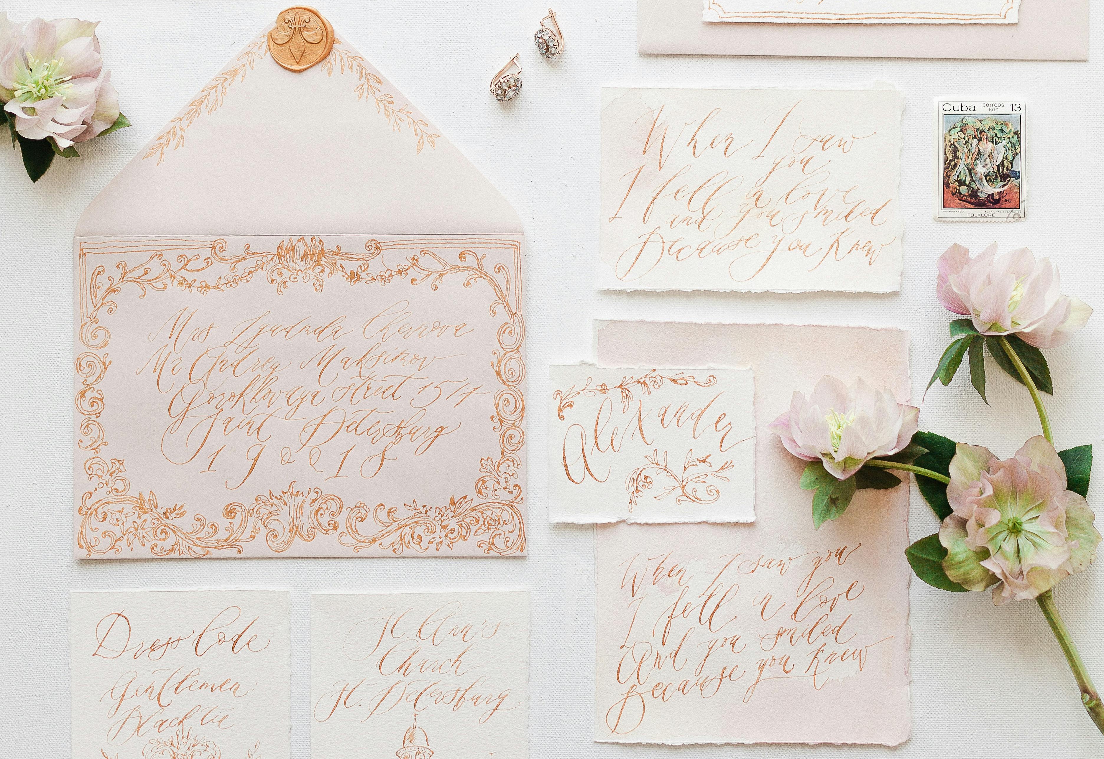 A wedding stationery flat-lay on blush and ivory paper with calligraphy writing and floral accents.