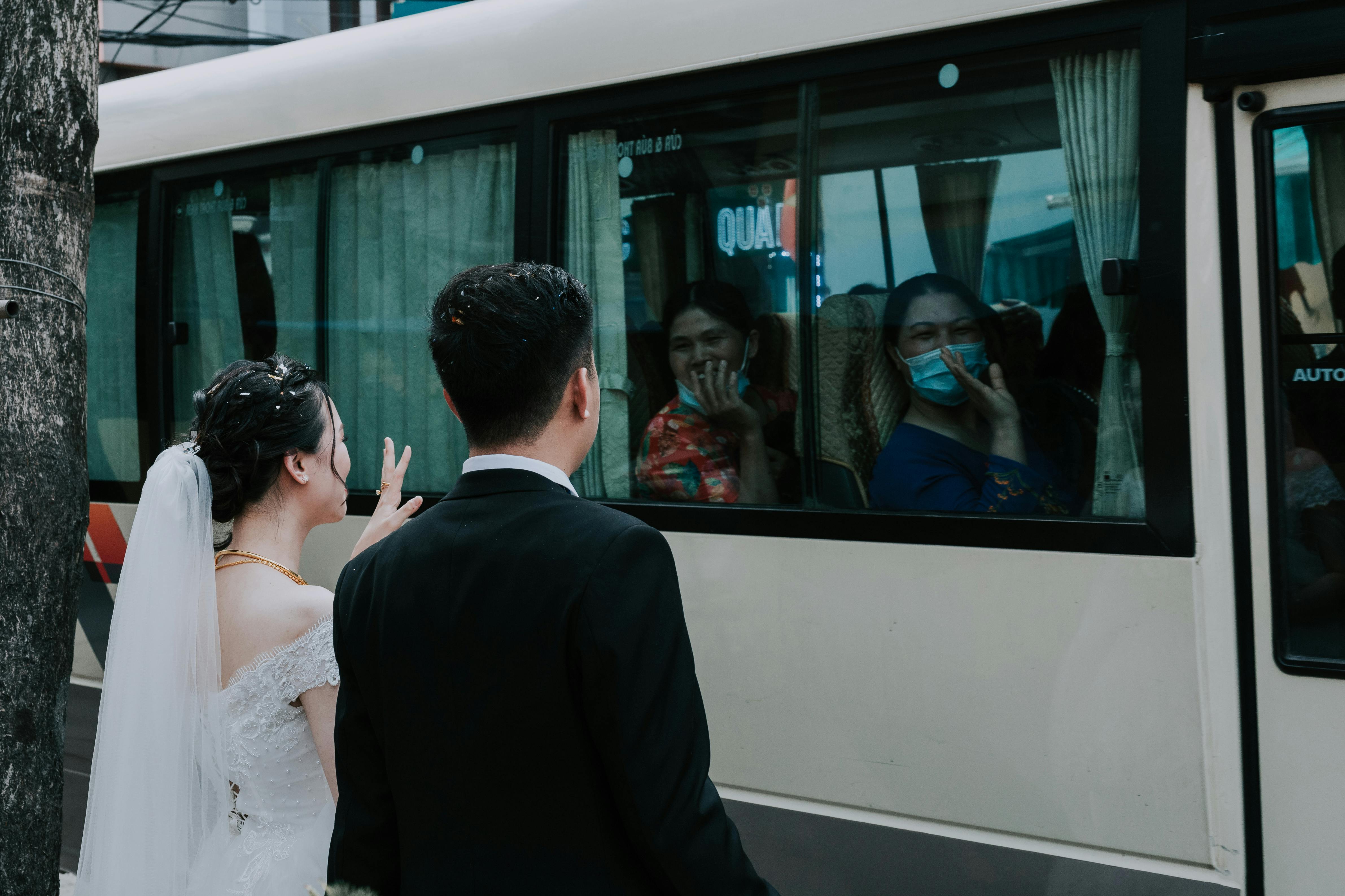 A bride and groom wave to their wedding guests, who are being transported on a bus.